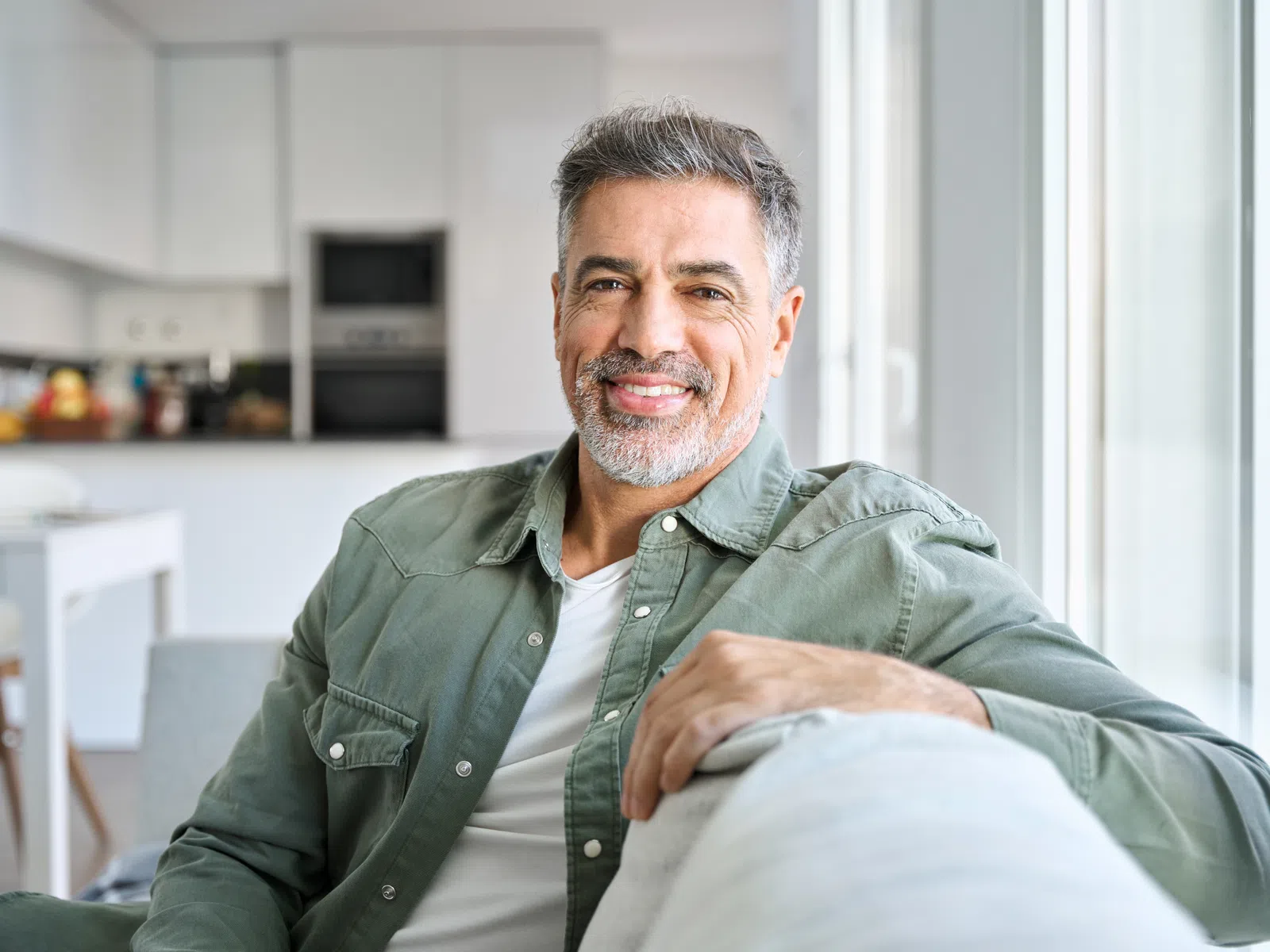 Smiling middle-aged man with gray hair sitting on a sofa in a bright modern kitchen and living room area.