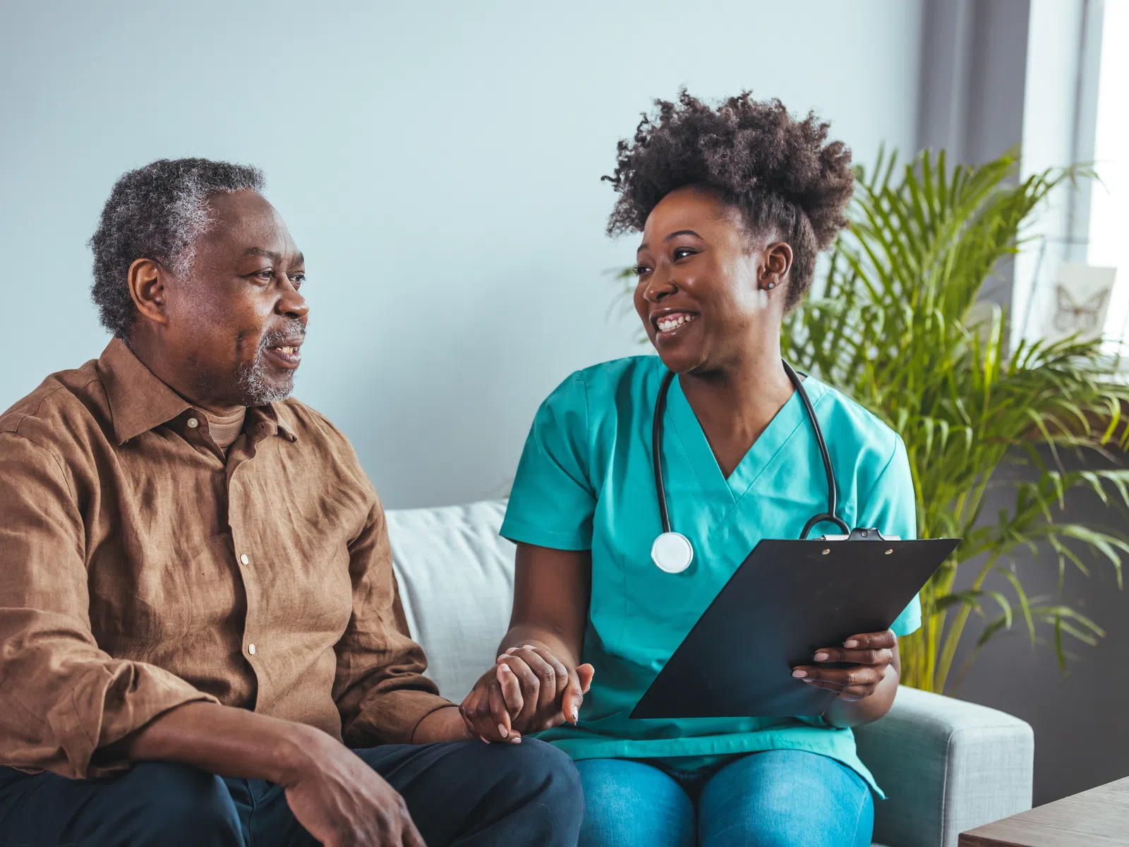 Smiling nurse in blue scrubs holds clipboard and patient’s hand during friendly consultation at home.