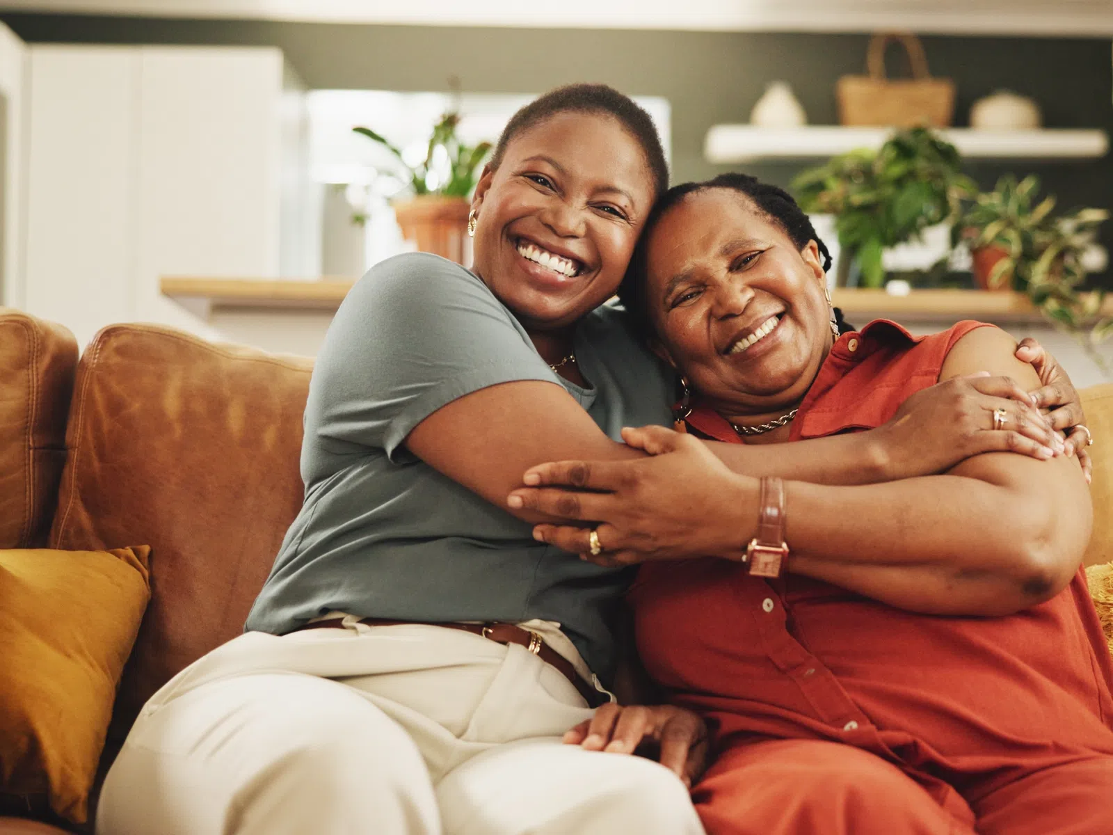 Two women smiling and hugging on a brown couch with yellow pillows in a cozy living room.