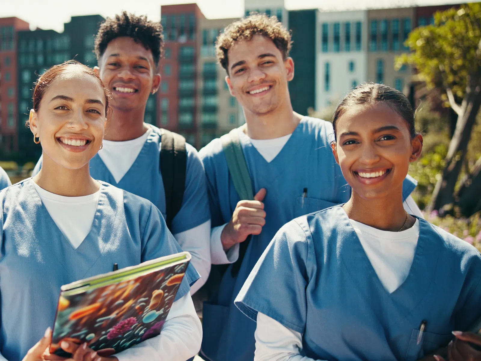 Diverse group of five smiling nursing students outdoors in blue scrubs holding books and tablets