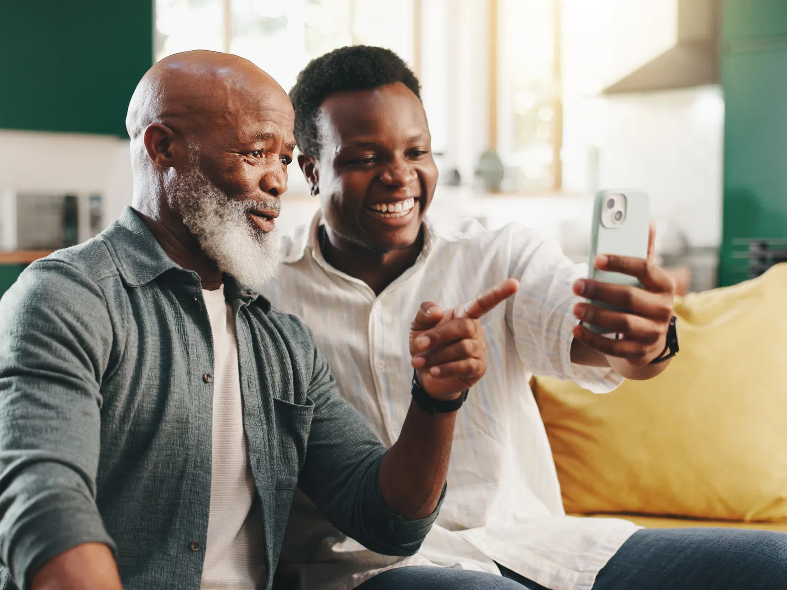 Smiling elderly man and young man taking a selfie together on a couch in a bright living room.