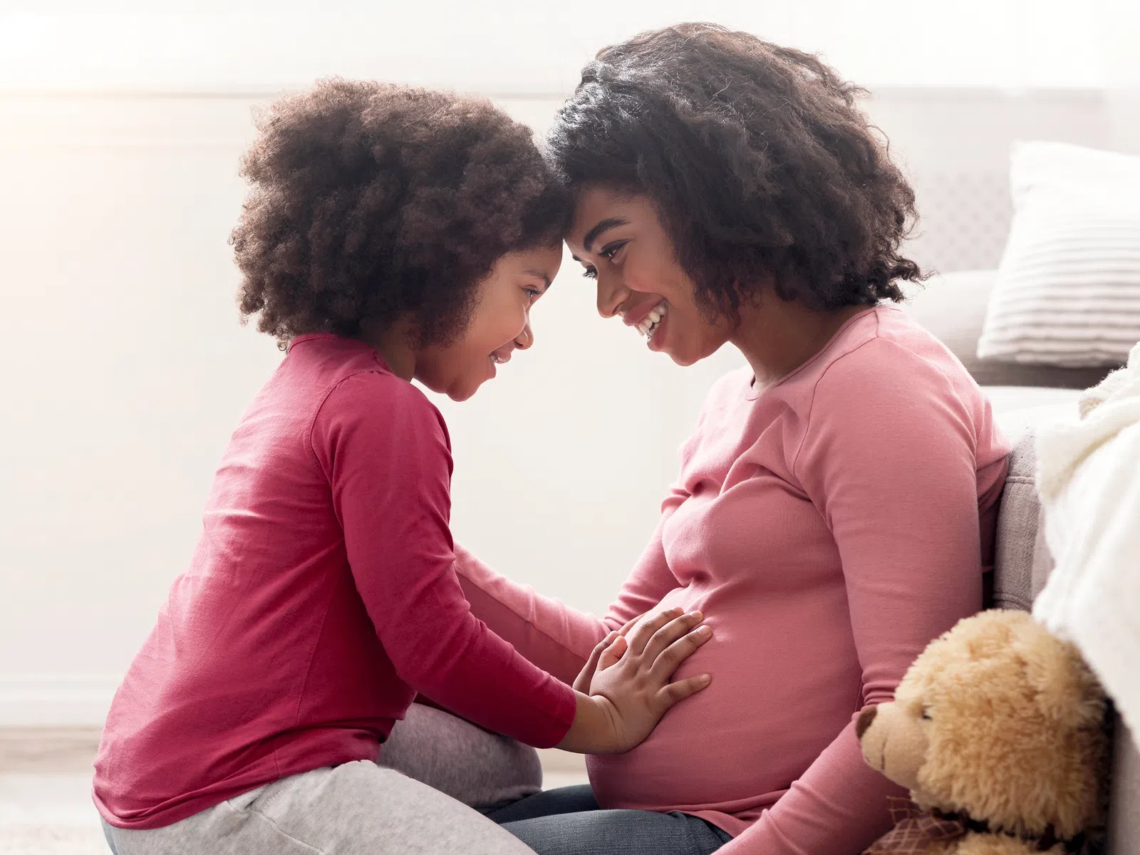 Pregnant woman and young girl smiling and touching baby bump in a cozy living room with teddy bear nearby