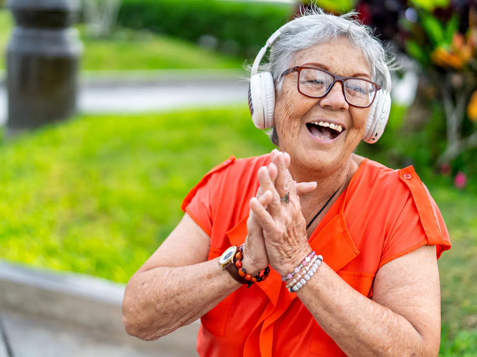 Happy elderly woman wearing headphones and an orange blouse enjoying music outdoors in a sunny garden.