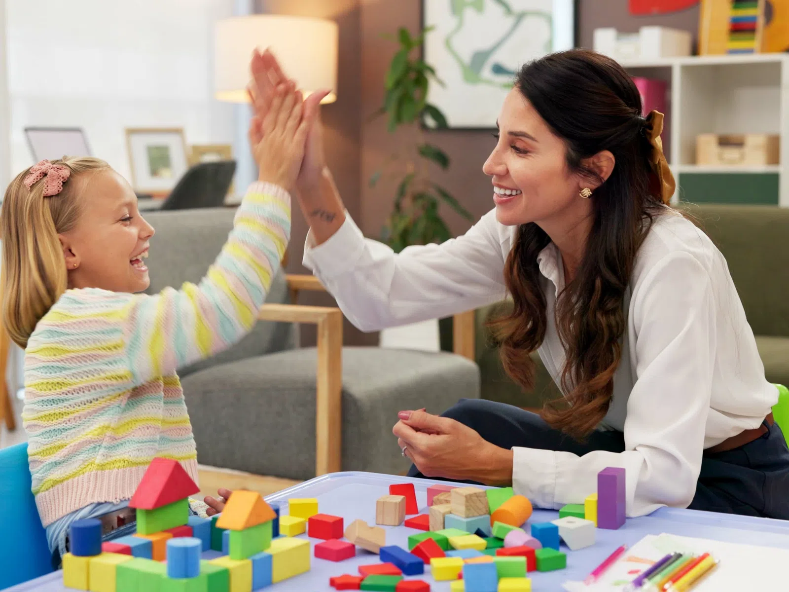 Happy woman and child giving high five while playing with colorful building blocks at table indoors