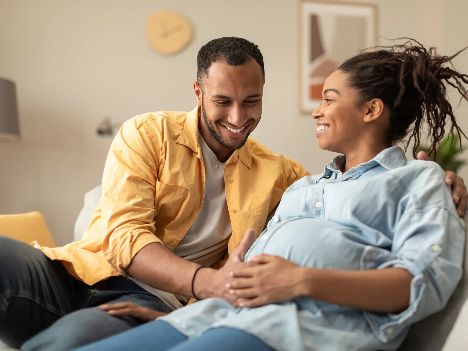 Happy couple sitting on couch, man touching pregnant woman's belly and both sharing a loving smile.