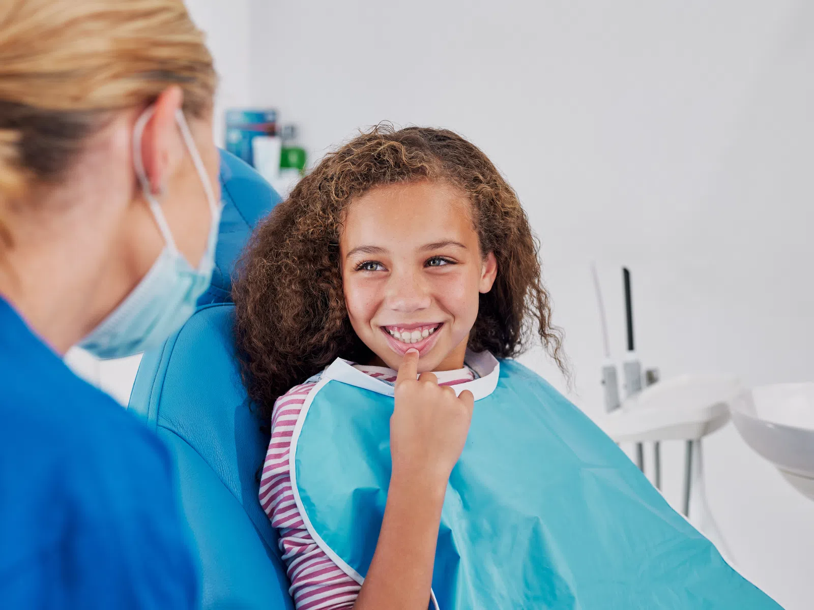 Smiling young girl sitting in a dental chair wearing a bib, interacting with a masked dental professional.
