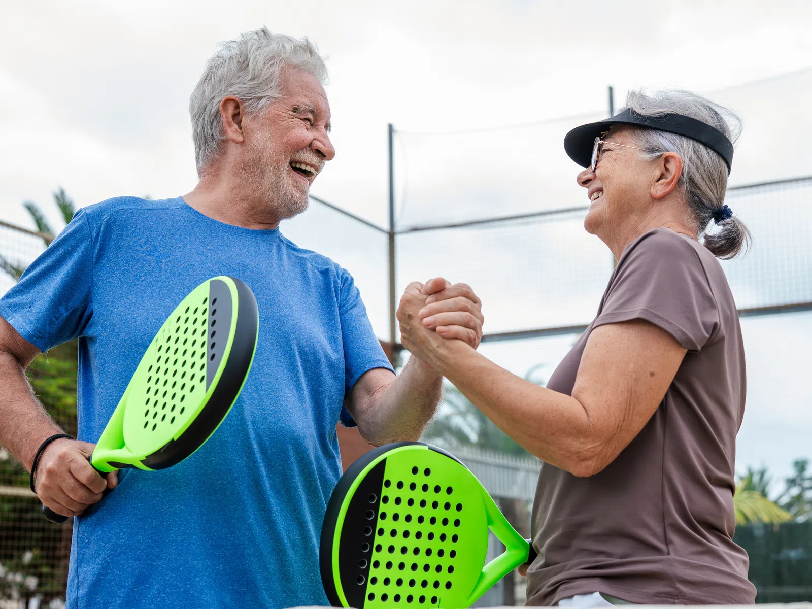 Senior couple enjoying a friendly paddle tennis match outdoors, smiling and shaking hands on the court.