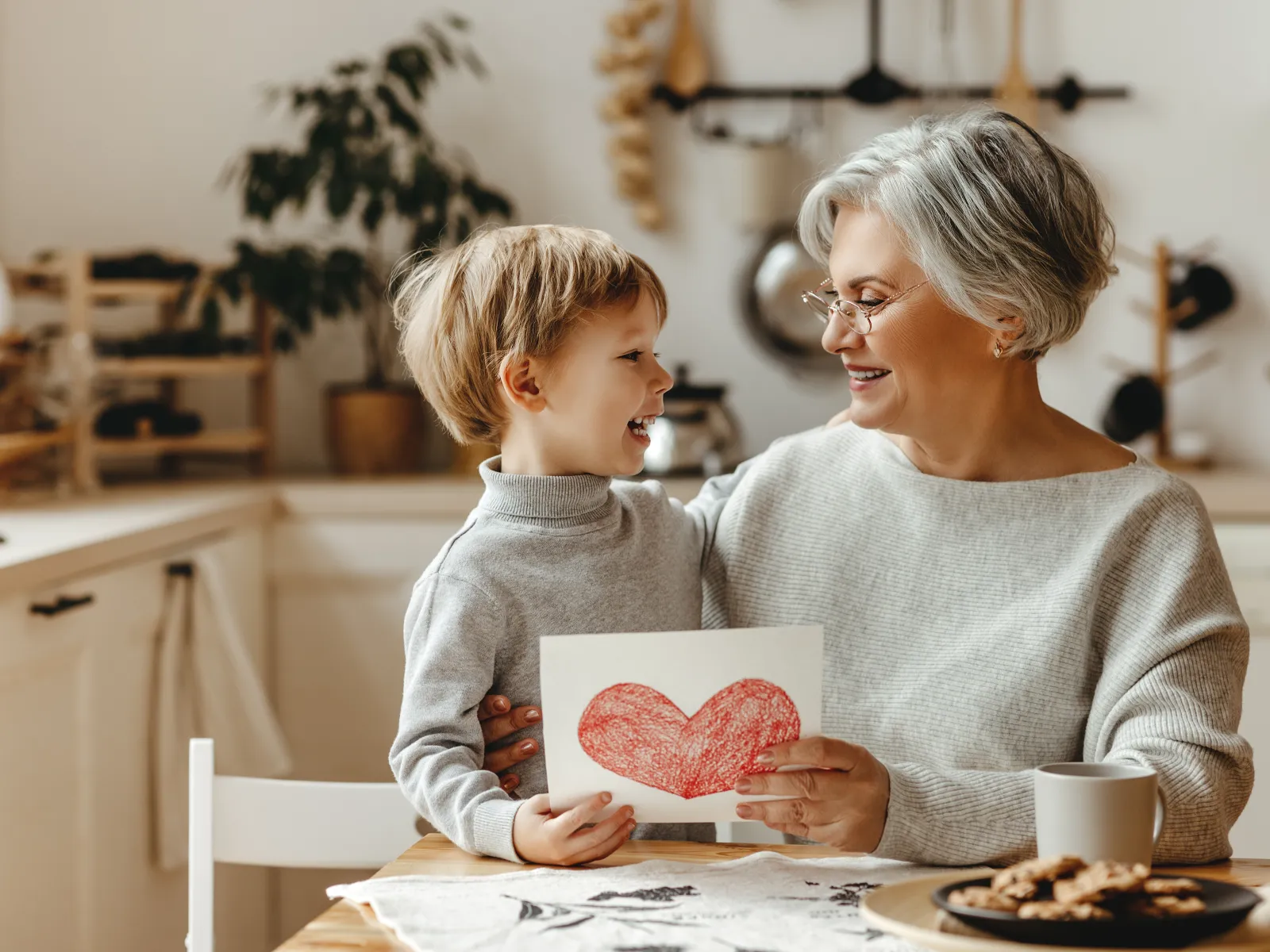 Smiling grandmother and grandson holding a handmade red heart drawing in cozy kitchen setting