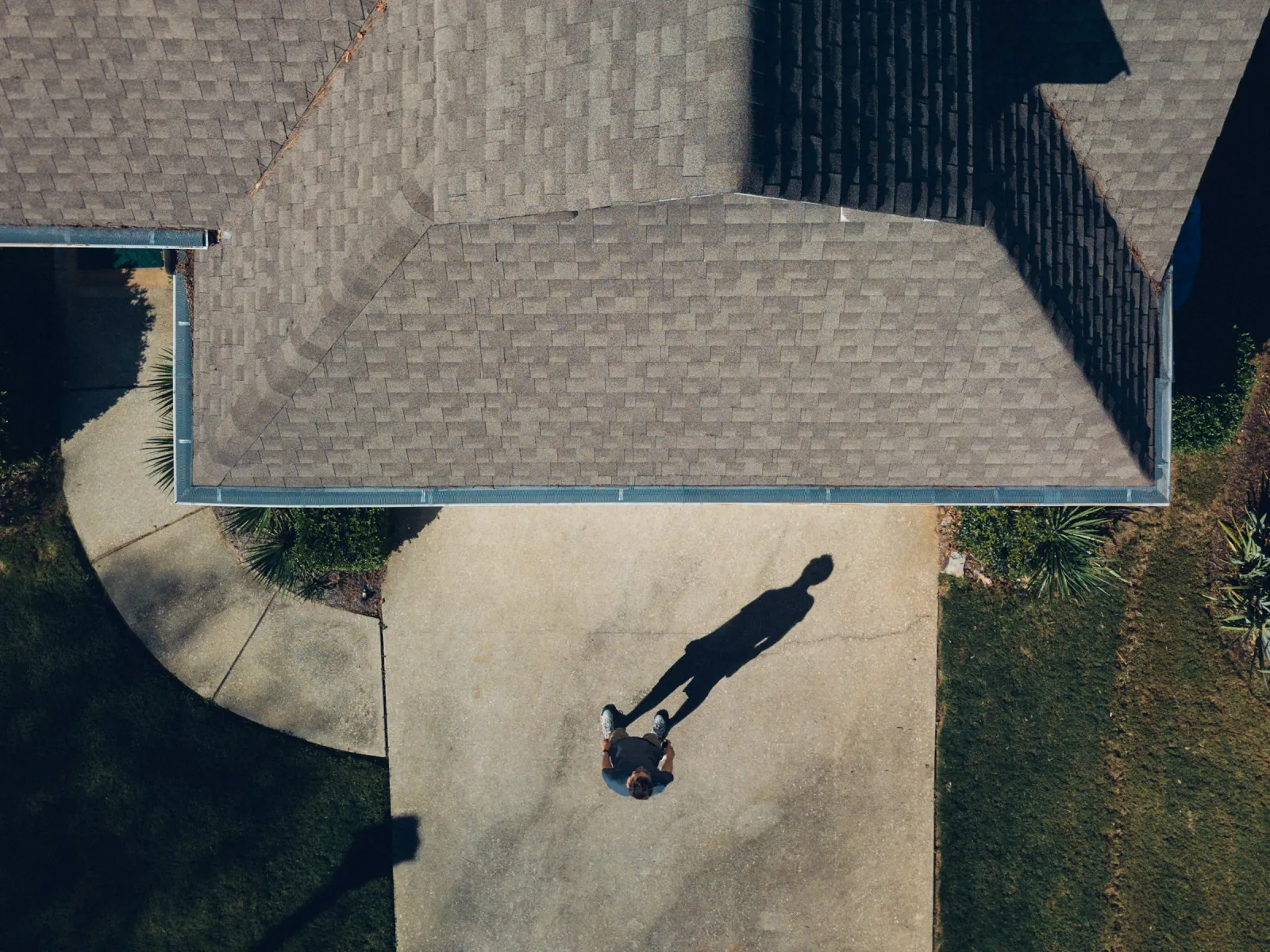 Aerial view of a person standing on a driveway casting a long shadow beside a house with gray shingle roof.