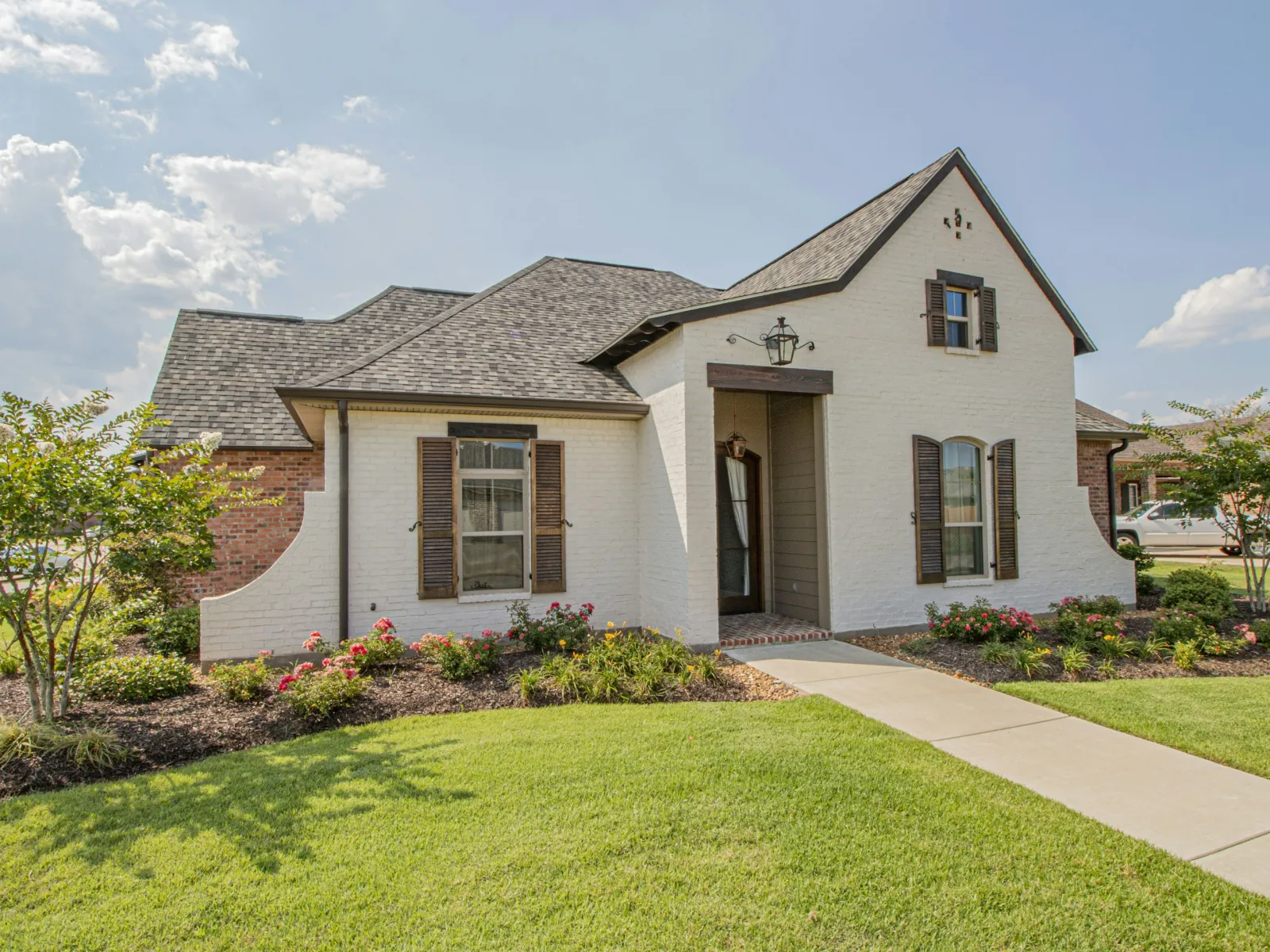 Single-story white brick house with dark shutters, manicured lawn, flower beds, and a concrete walkway under a blue sky.