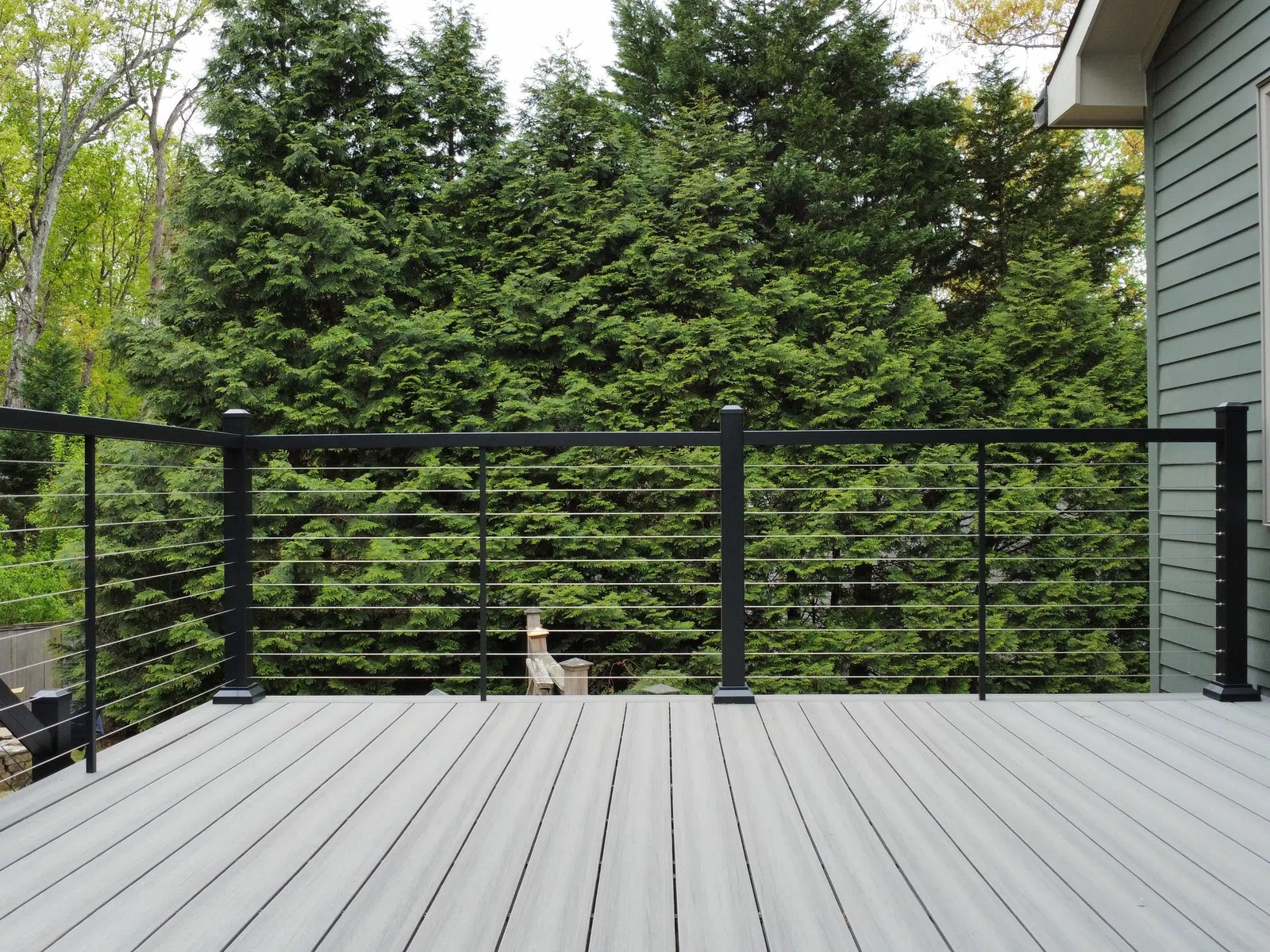 Modern gray outdoor deck with black metal railing overlooking dense green trees outside a gray house.