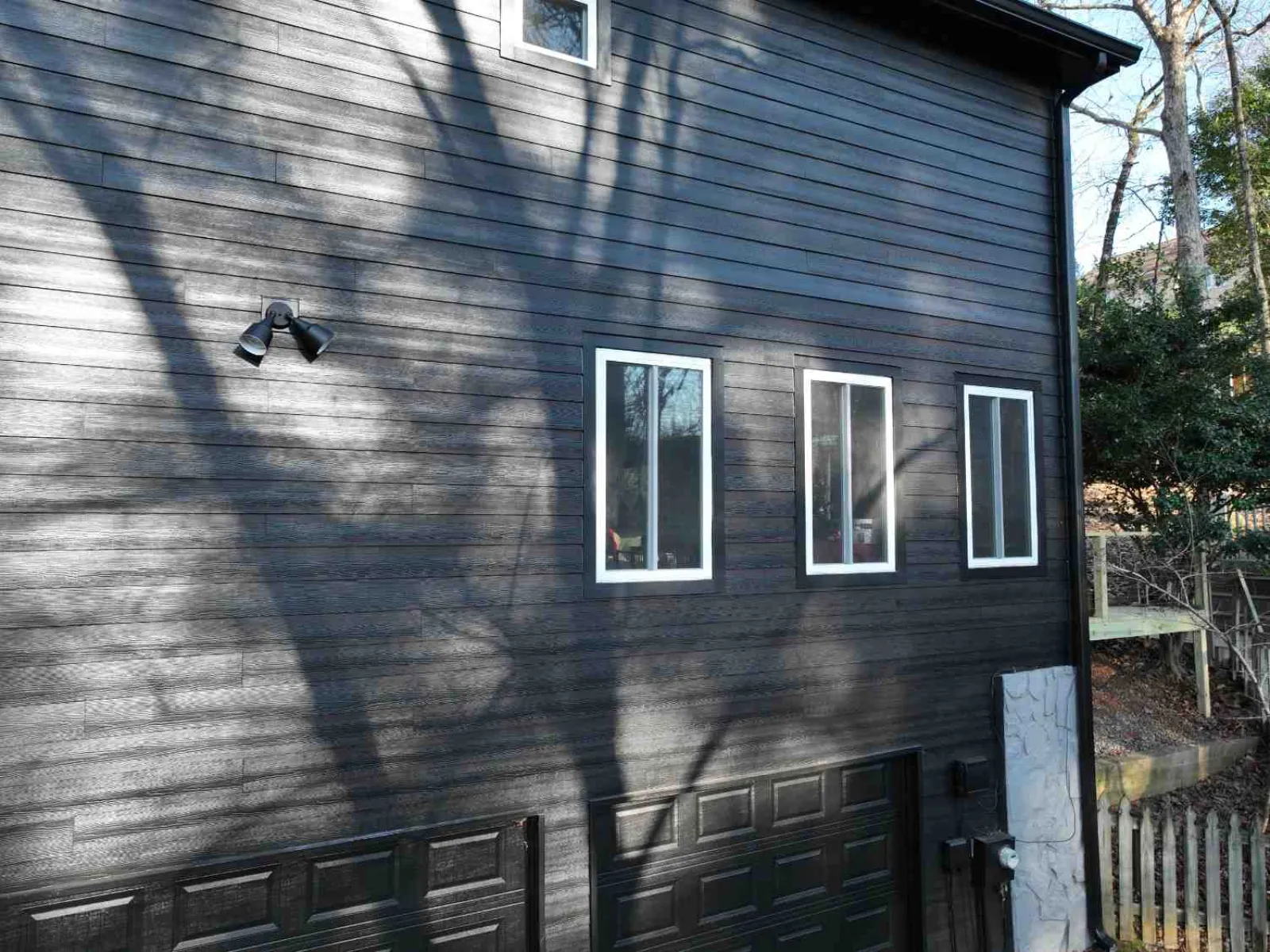 Exterior side of a modern black house with three white-trimmed windows and shadows of tree branches.