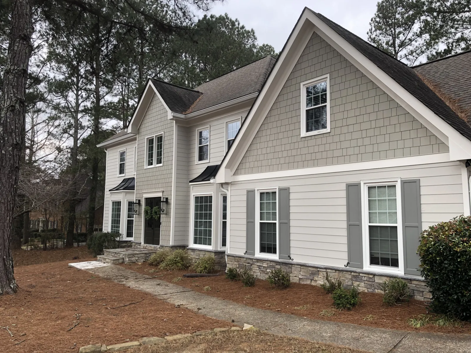 Large two-story suburban house with gray siding, white trim, stone foundation, and surrounding pine trees.