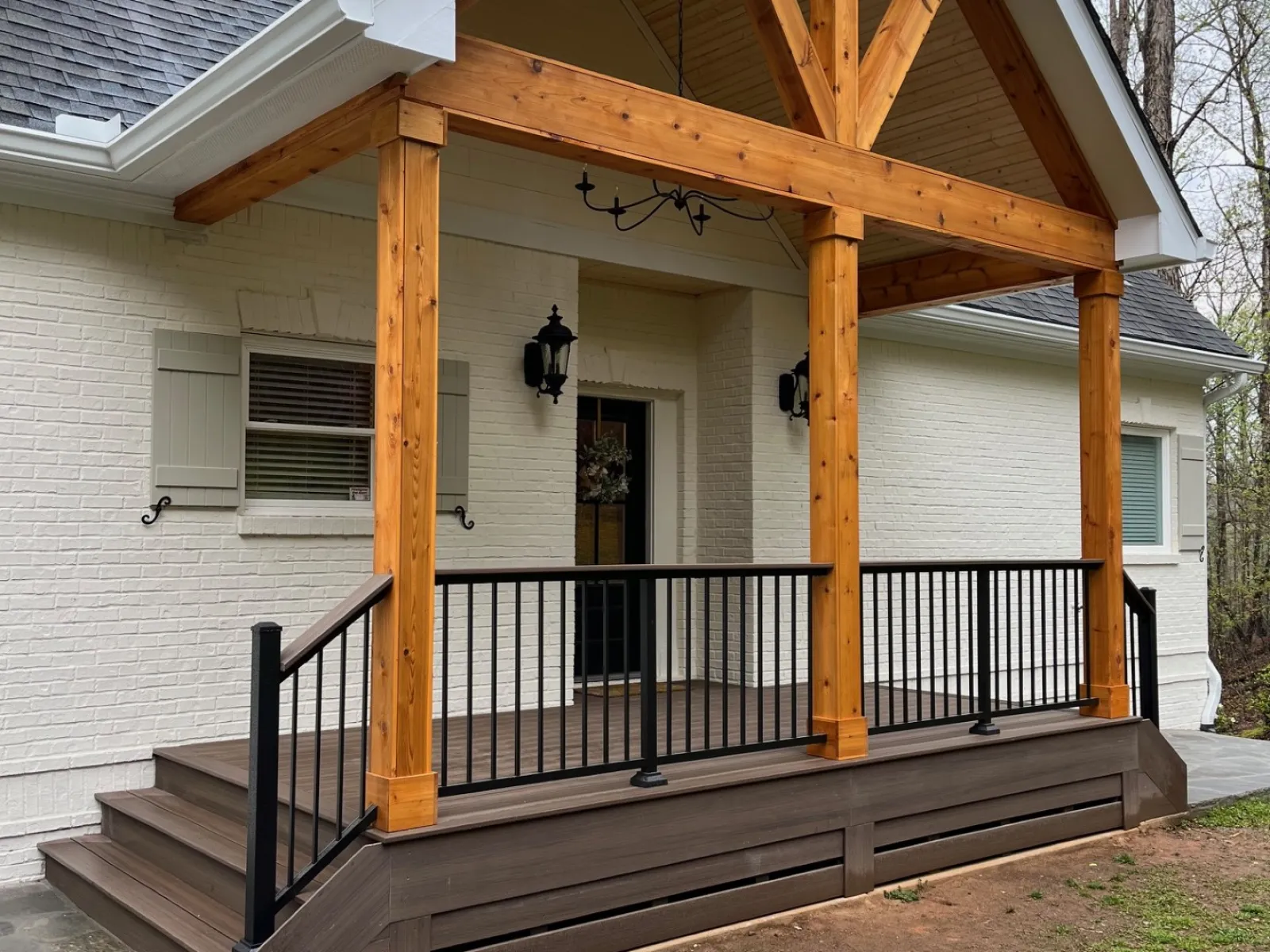 Covered front porch with wooden beams, black railing, and steps leading to a white brick house entrance.