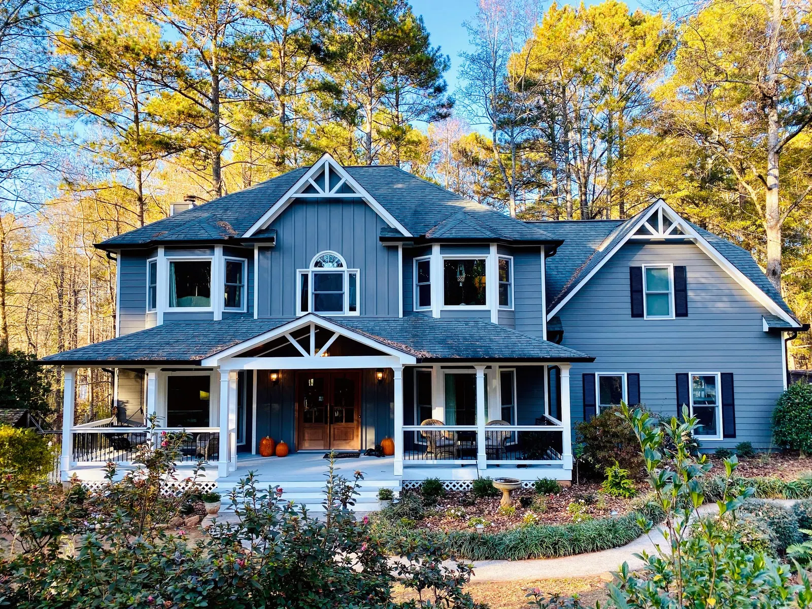 Large two-story blue house with white trim surrounded by autumn trees and a landscaped yard with a curved pathway.