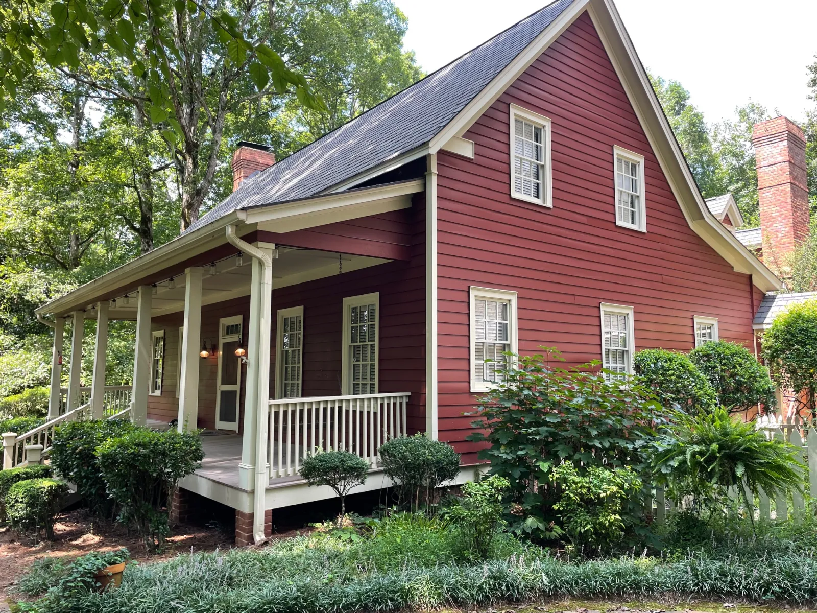 Red wooden house with white trim, front porch, surrounded by green shrubs and trees on a sunny day
