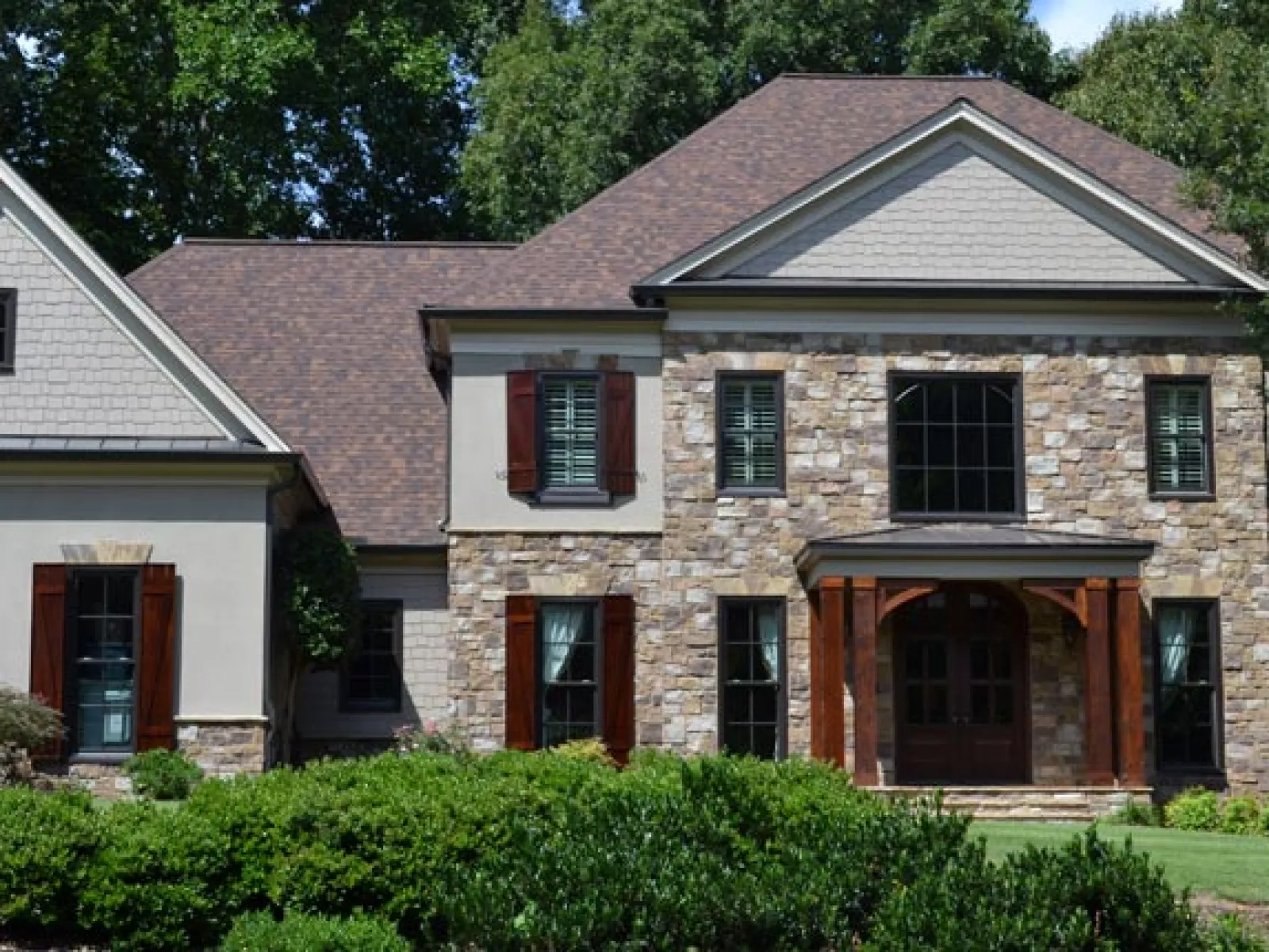 Elegant two-story house with stone facade, landscaped garden, and bright windows surrounded by trees.
