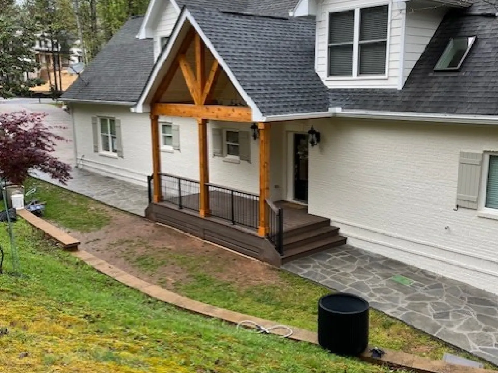White house with a black roof, wooden porch beams, stone walkway, and sloped grassy yard with trees.