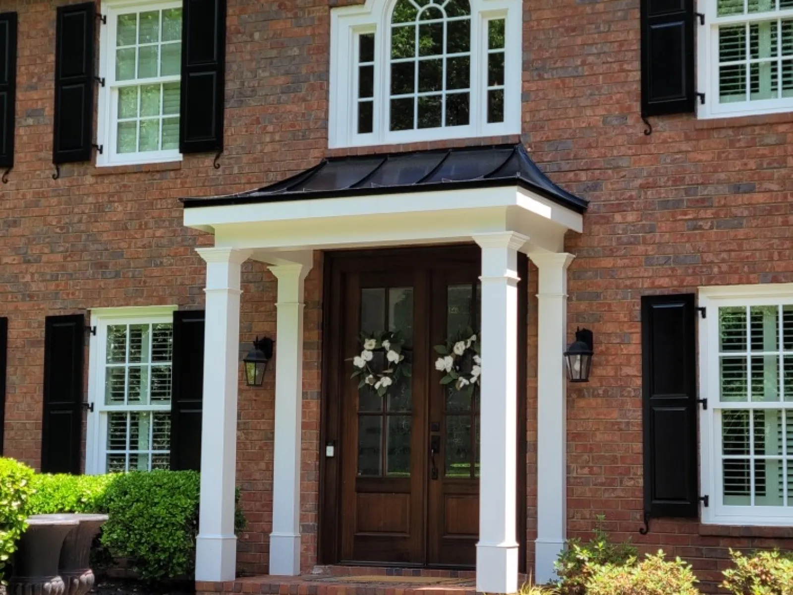 Brick house entrance with dark wooden double doors, white columns, black shutters, and greenery.
