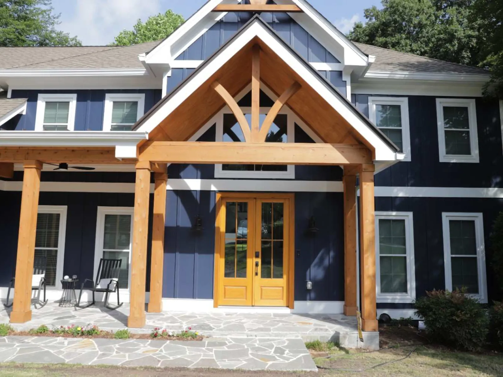 Modern blue two-story house with wooden porch, stone pathway, and glass double front doors under a triangular roof.