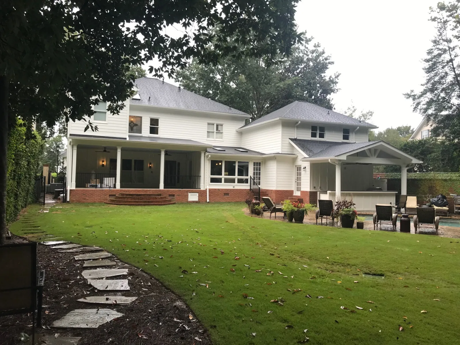 Large white two-story house with covered patio, manicured lawn, stone path, and outdoor seating area near pool.