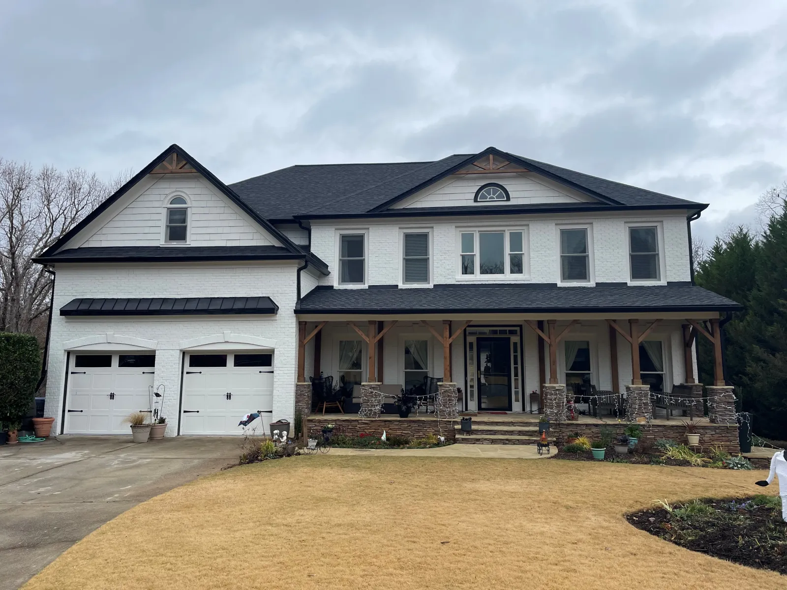 Two-story white house with black roof, large front porch, two-car garage, and a dry lawn under cloudy sky