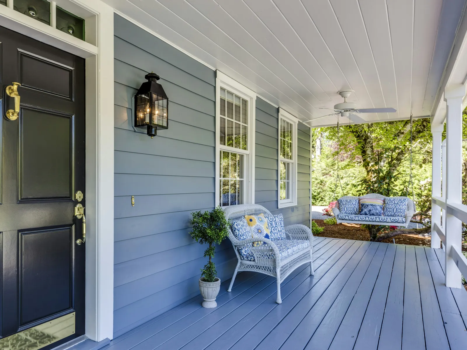 Cozy blue front porch with white wicker bench, swing, potted plant, black door, and outdoor lantern light.