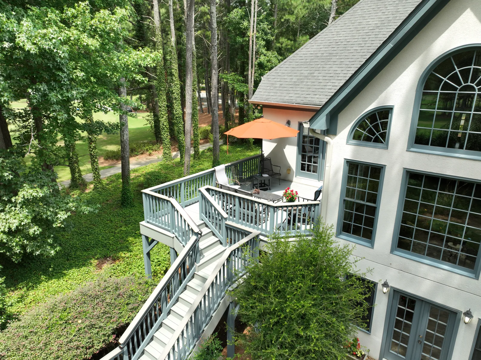 Modern white house with large windows, blue deck railing, outdoor seating, and surrounding green trees and lawn.