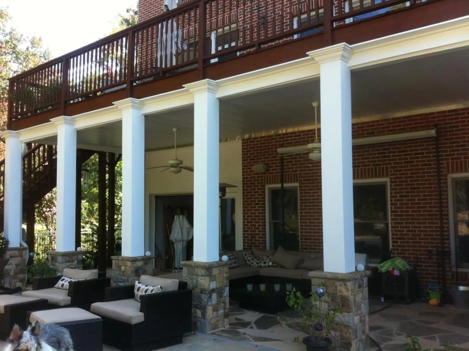 Outdoor patio area with stone pillars, white columns, ceiling fans, and cushioned seating under a wooden deck.