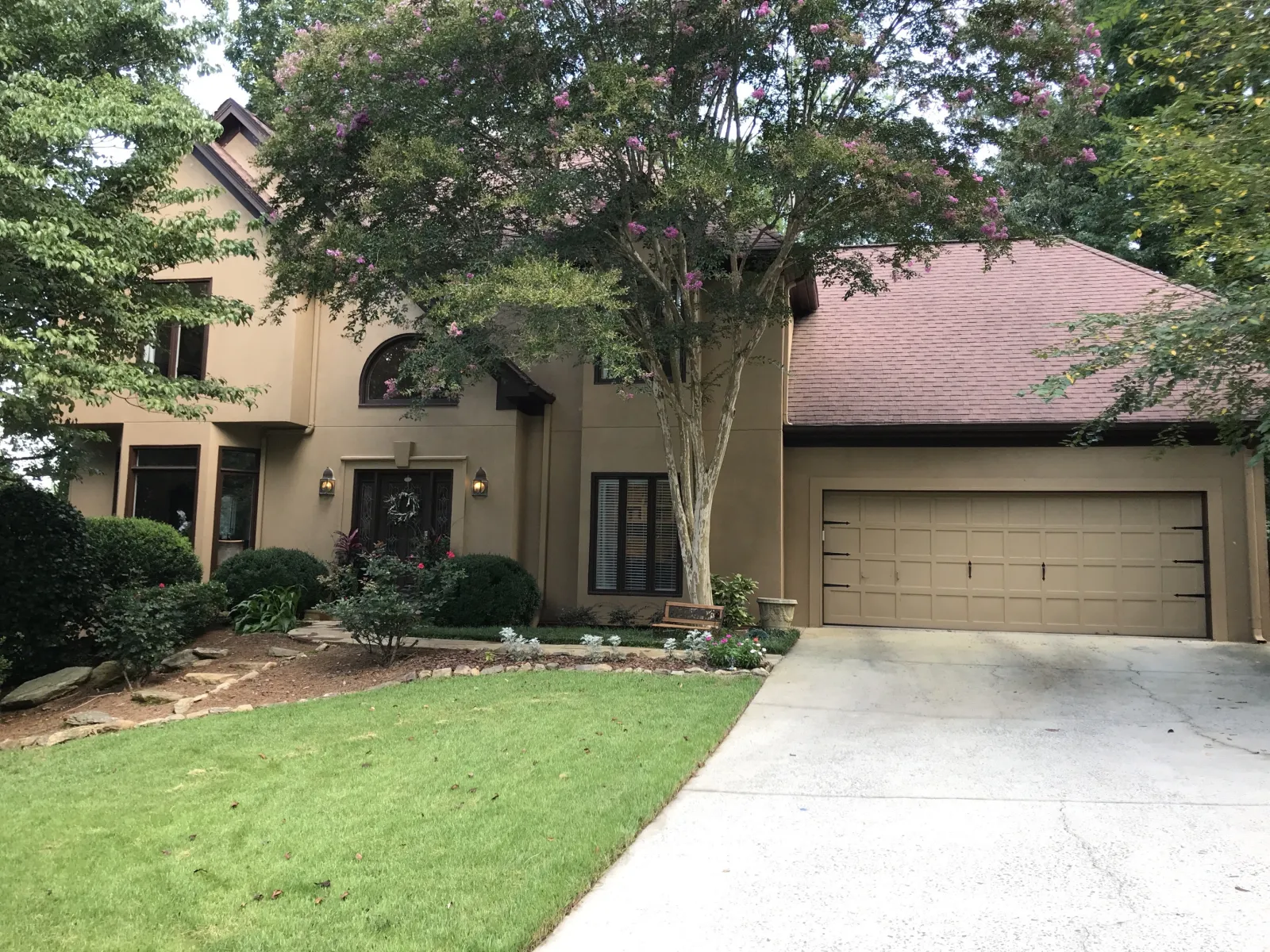Two-story beige house with a brown roof, garage, front yard, and flowering trees in a suburban neighborhood.
