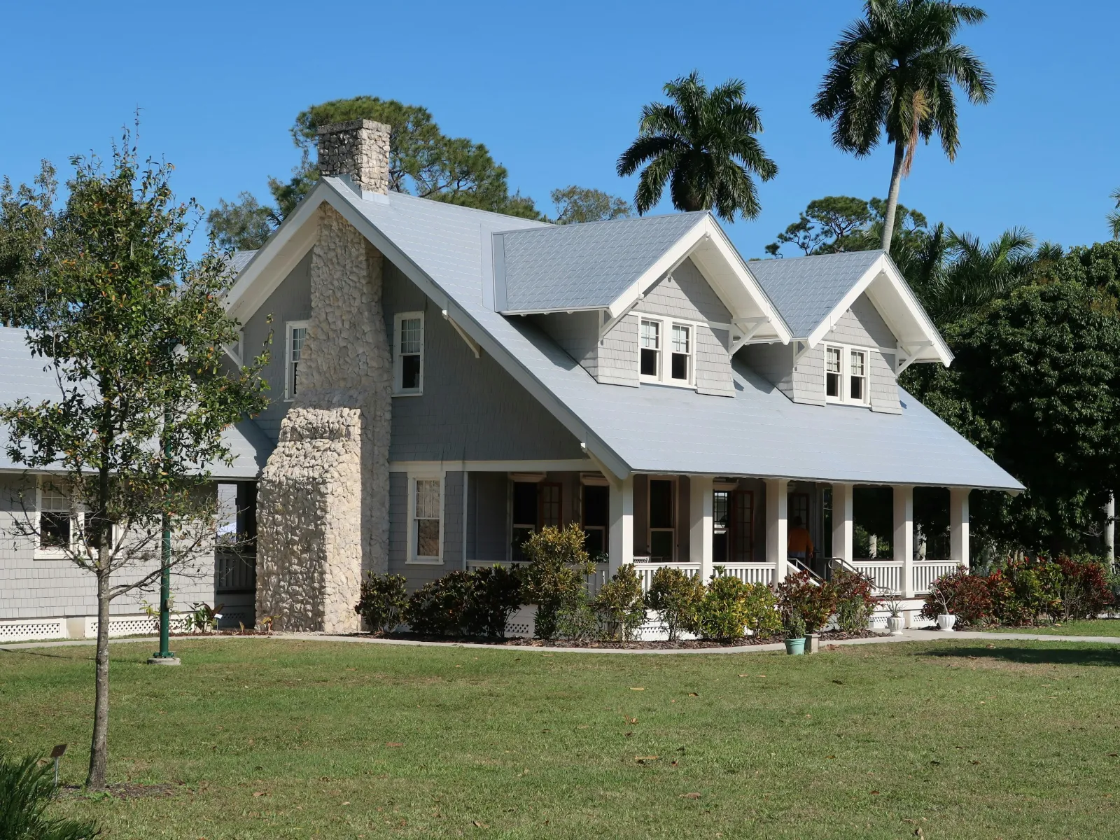 Gray craftsman-style house with stone chimney, large porch, and lush green lawn under blue sky