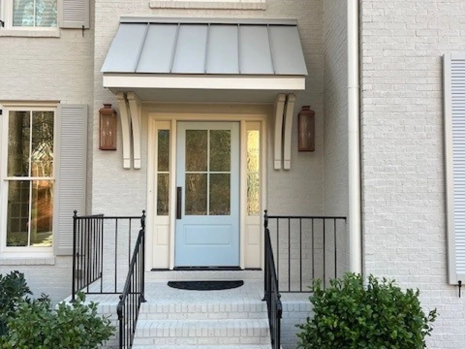 Front entry of white brick house with pale blue door, black railing, steps, and greenery on both sides.