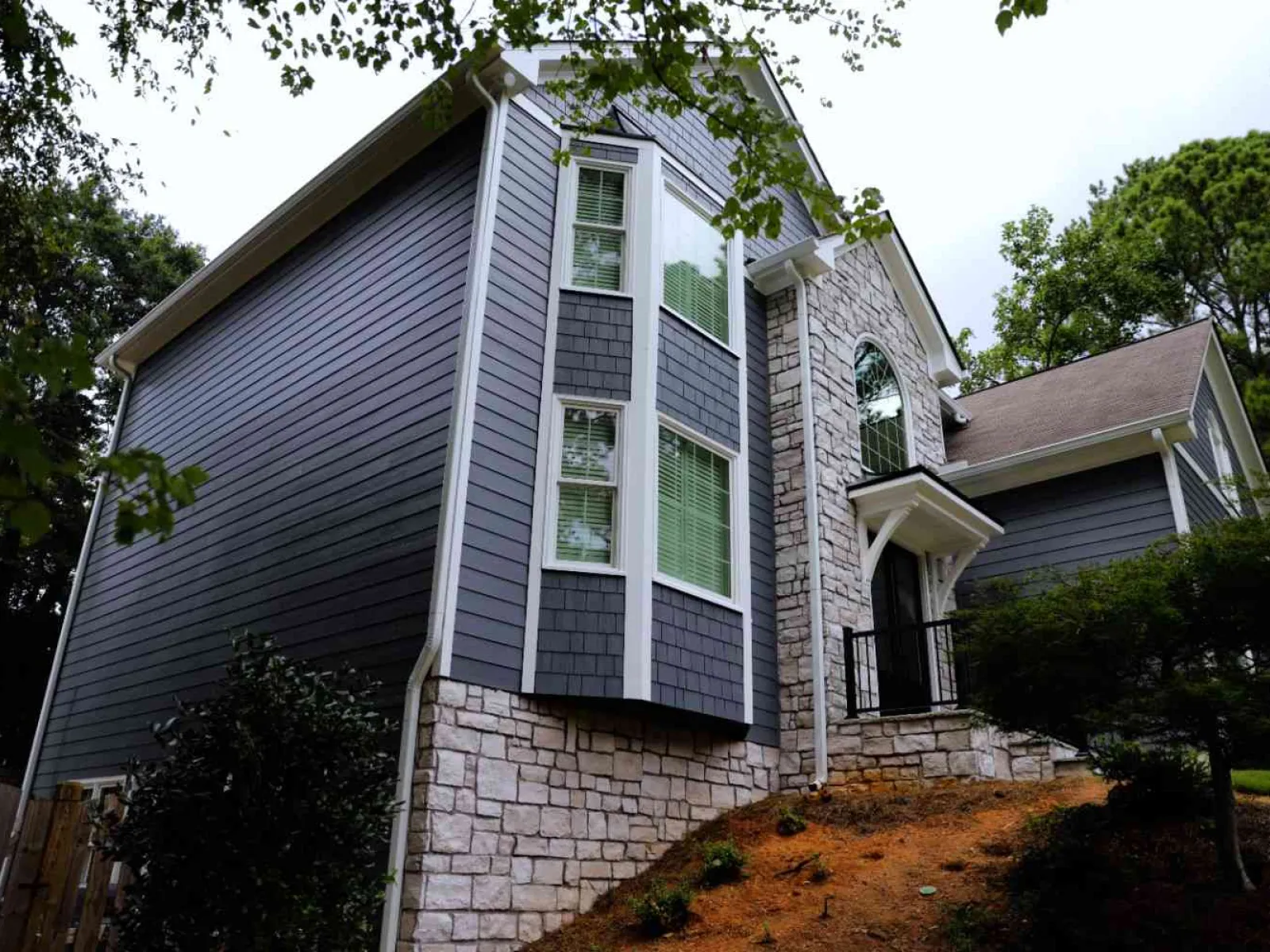 Modern two-story house with gray siding and stone facade surrounded by trees and shrubs on sloped land