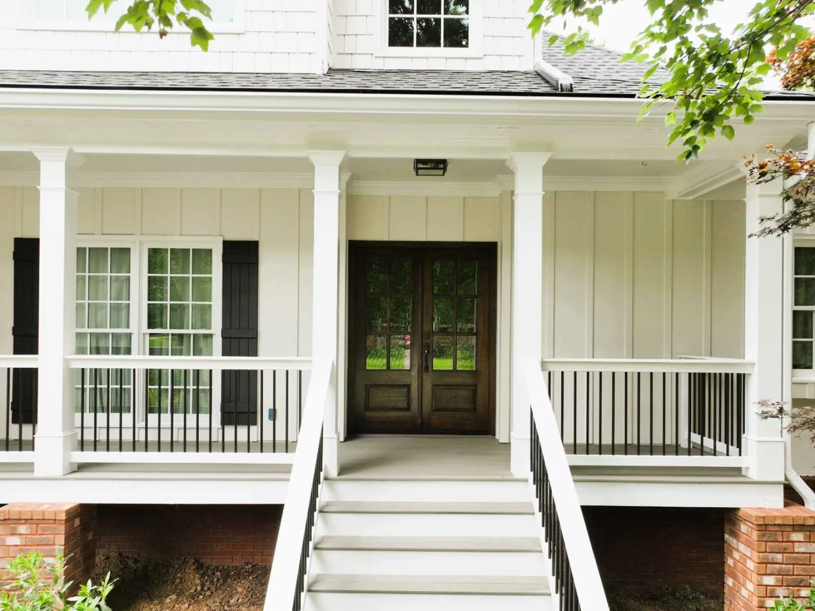 Front porch of a white house with wooden double doors, black railings, and brick foundation.
