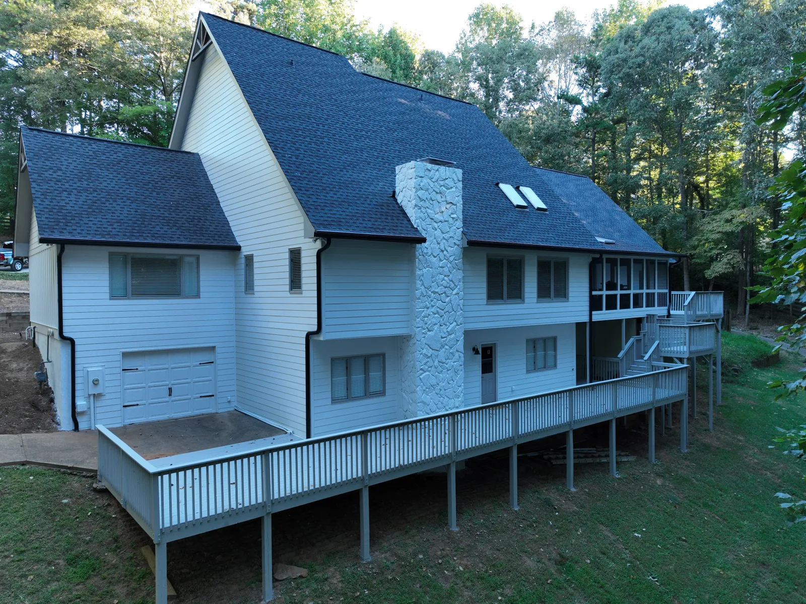 White multi-level house with black roof, stone chimney, wraparound deck, surrounded by trees on sloped land.