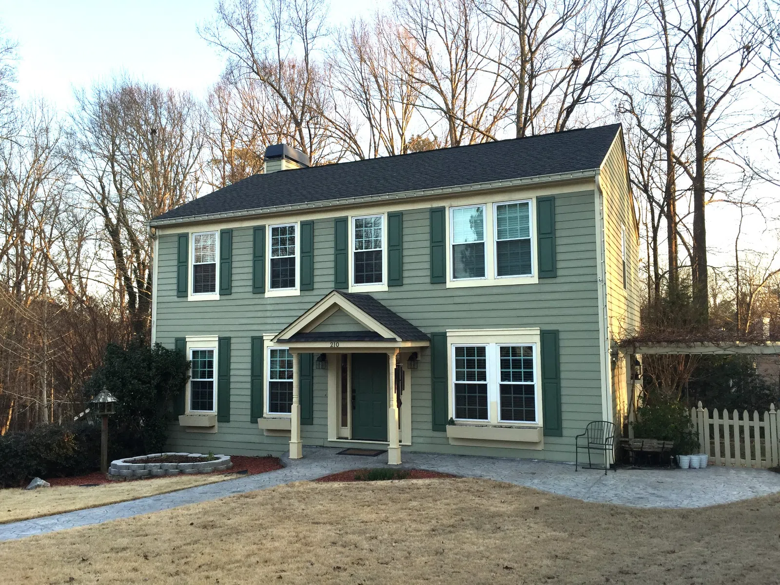 Two-story green house with white trim, surrounded by leafless trees and a dry front yard in winter.