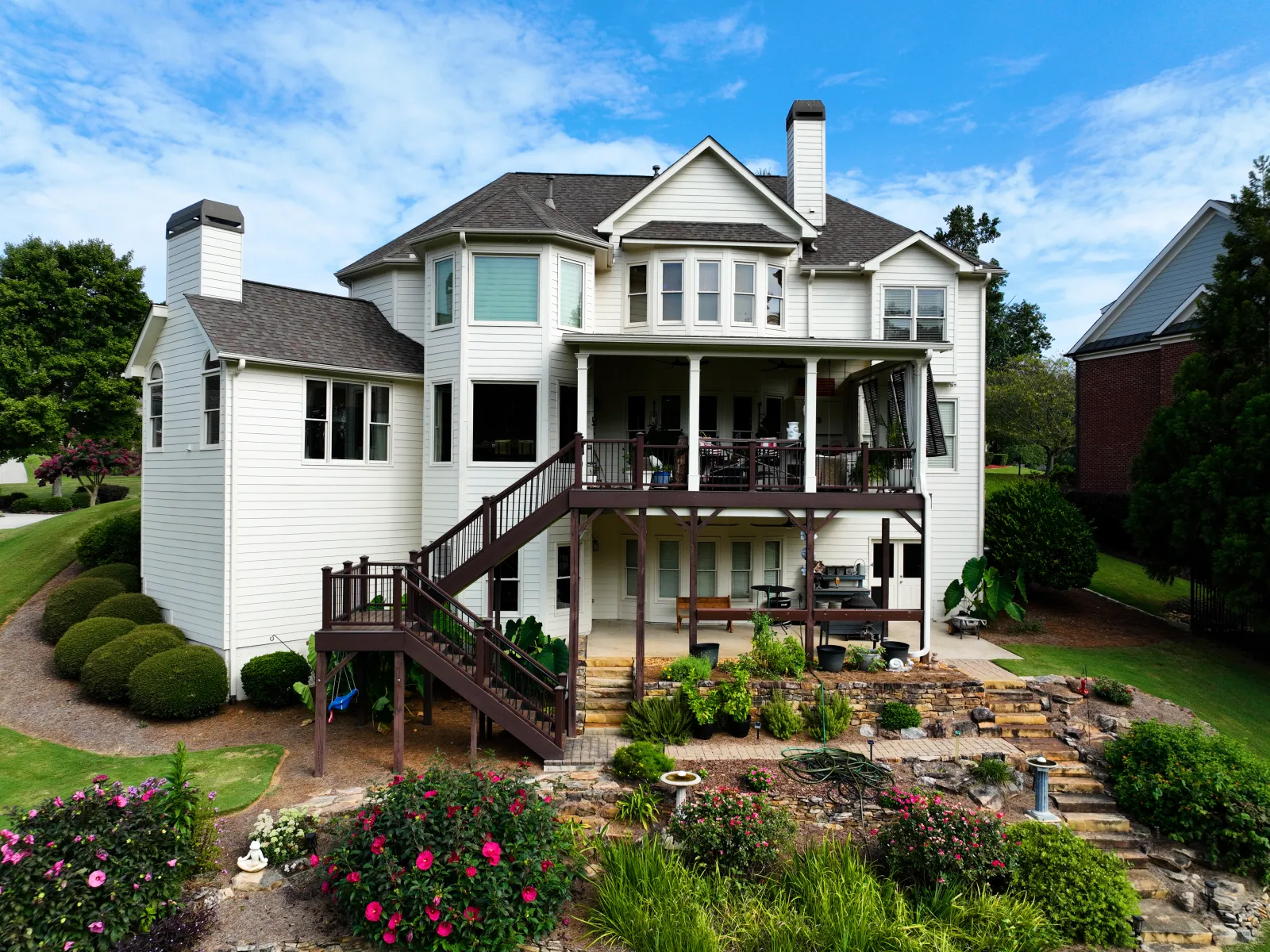 Large white two-story house with a raised wooden deck, garden, and blue sky with clouds.