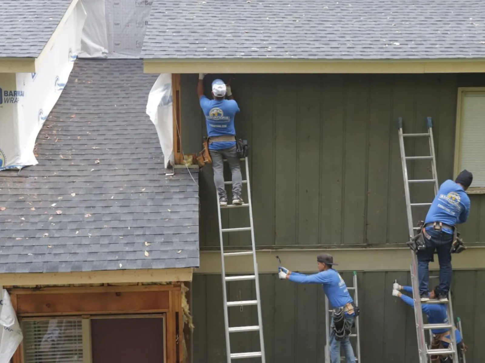 Construction workers install siding and wrap on a two-story house using ladders under a gray roof.