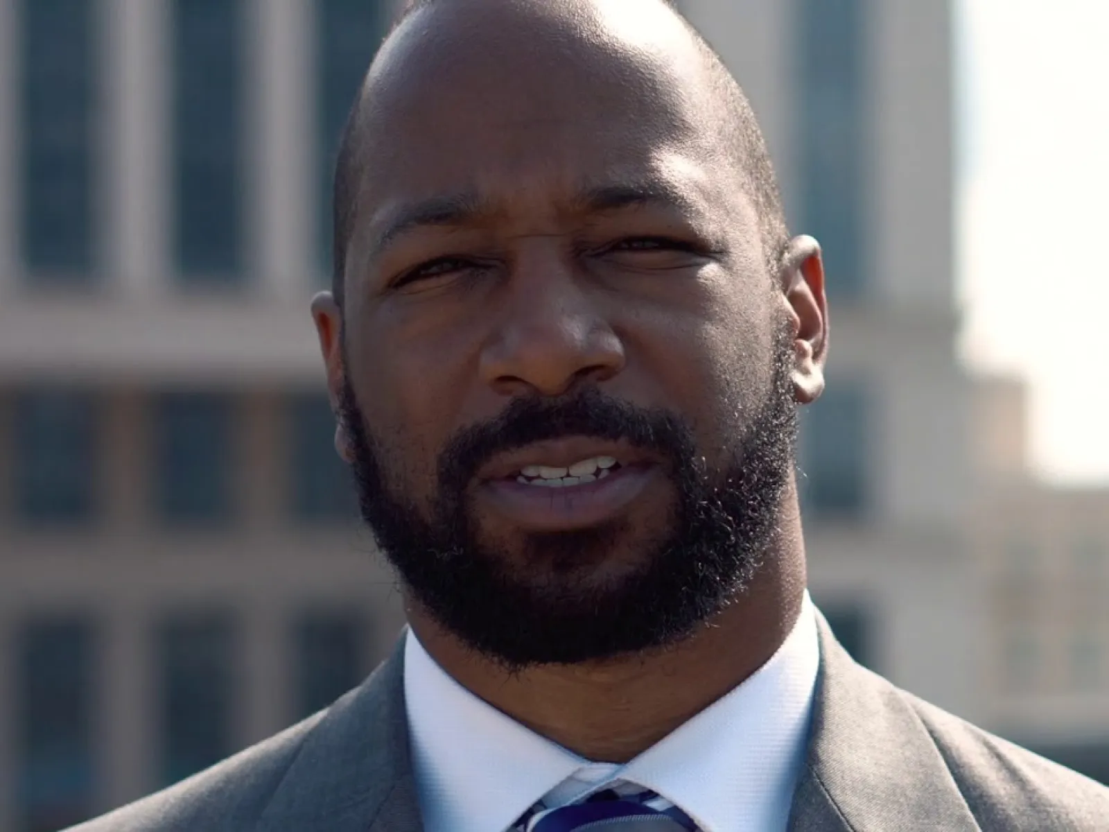 Confident bald man with beard wearing a suit and tie outdoors in front of modern buildings on a sunny day
