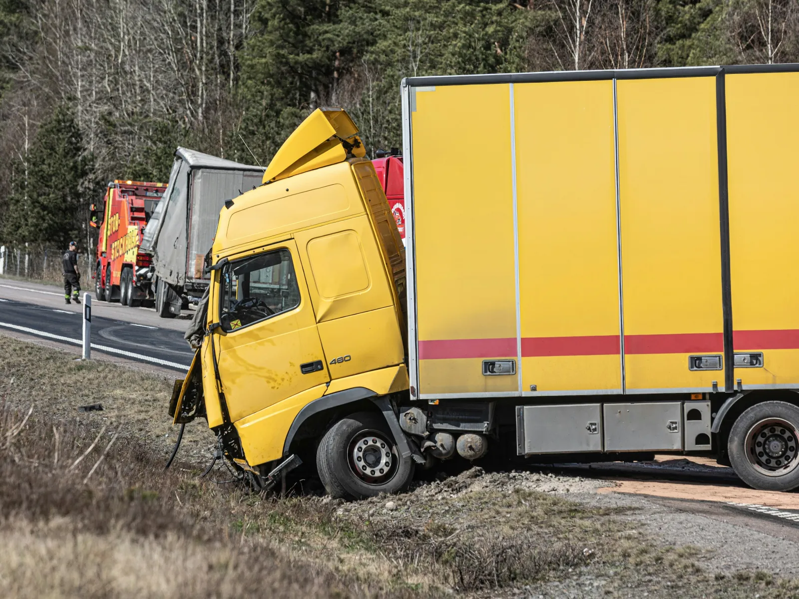 Yellow delivery truck crashed off the road with damaged front near a highway surrounded by trees and emergency vehicles.