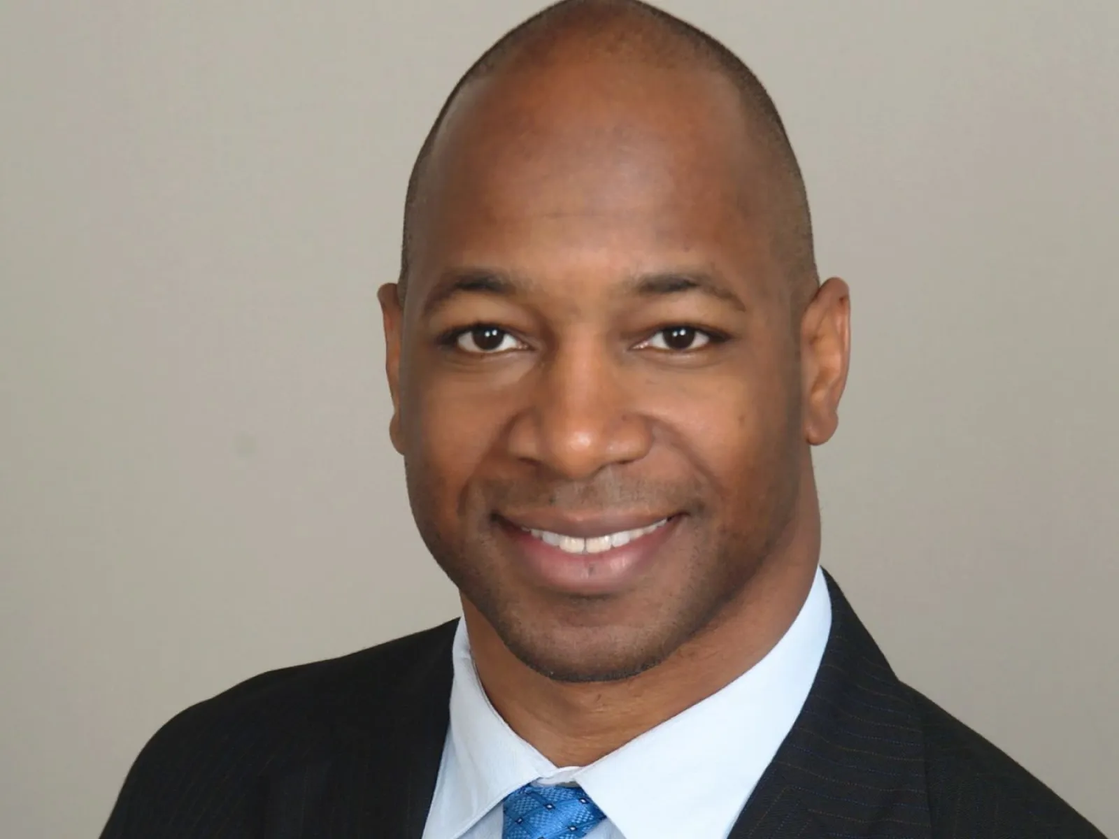 Smiling African American man in a dark suit with a blue patterned tie against a plain background