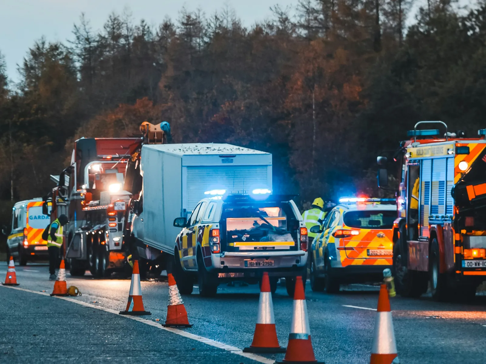 Emergency vehicles with flashing lights attend a roadside incident at dusk with safety cones and personnel present.
