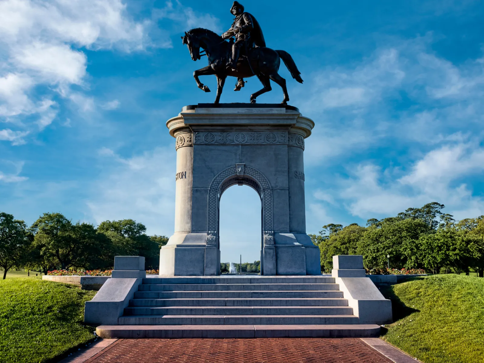 Statue of a mounted figure against a blue sky, surrounded by green park landscapes.