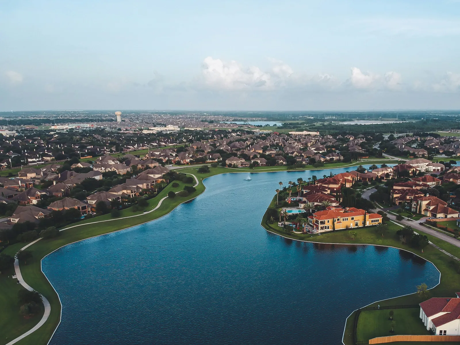 Aerial view of suburban neighborhood with large lake, waterfront homes, green spaces, and distant urban skyline.