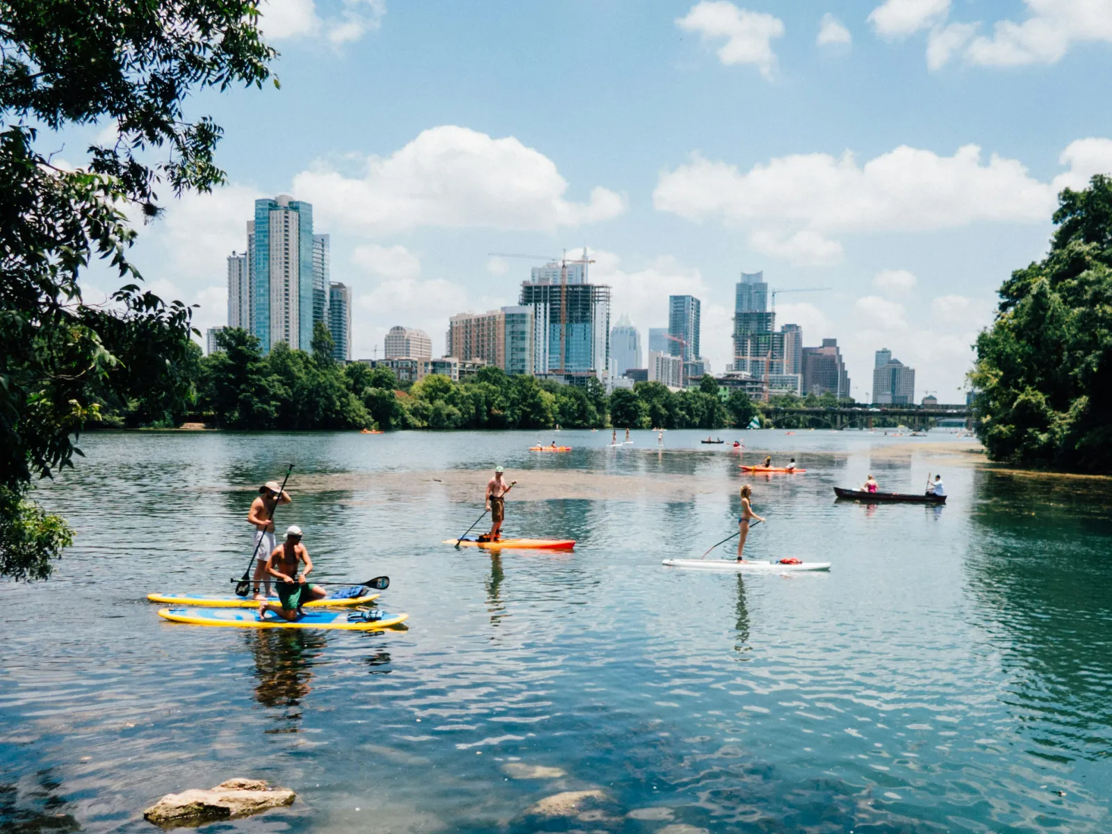 People paddleboarding on a calm lake with city skyline and green trees in the background.