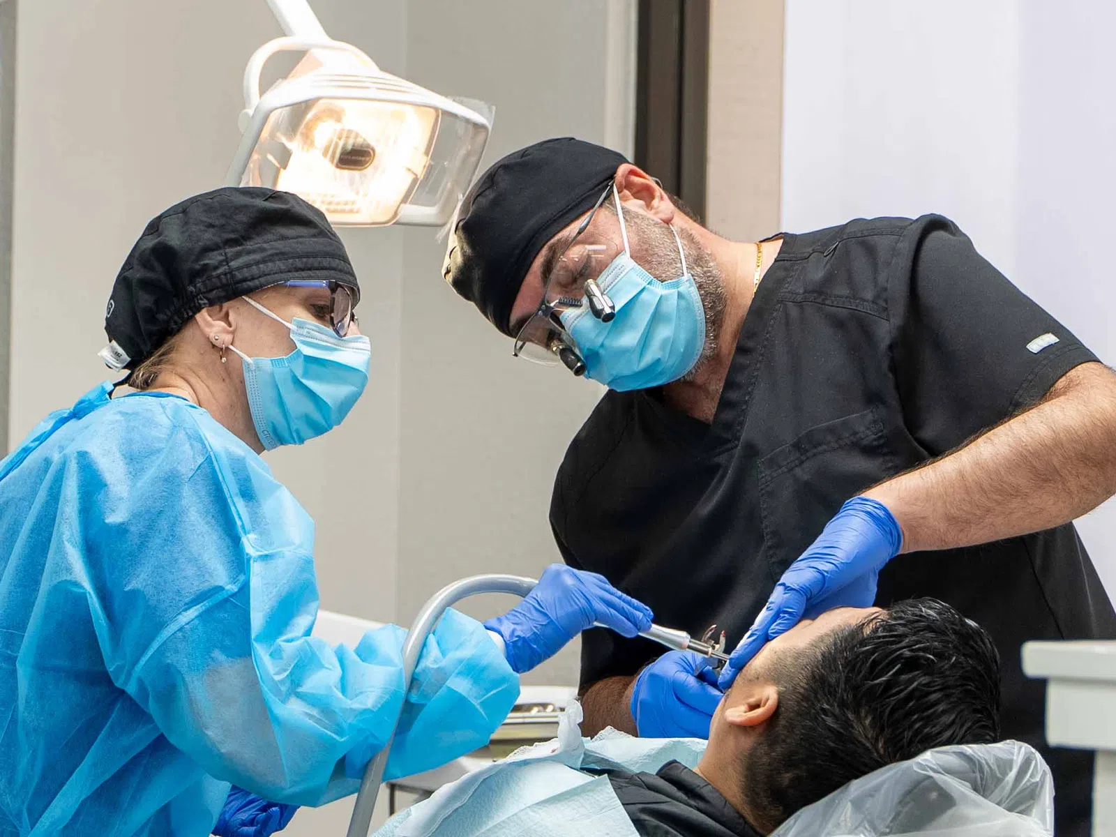 Dentists in masks and gloves treating a male patient in a modern dental clinic under bright light.