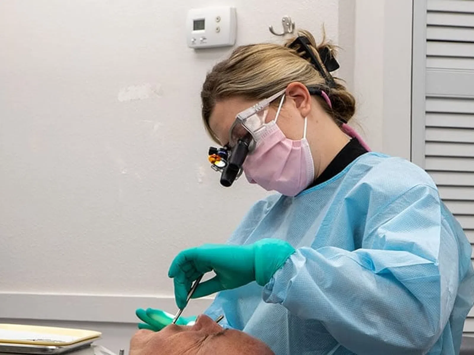 Dentist in blue scrubs and magnifying glasses performs dental work on a patient with green gloves