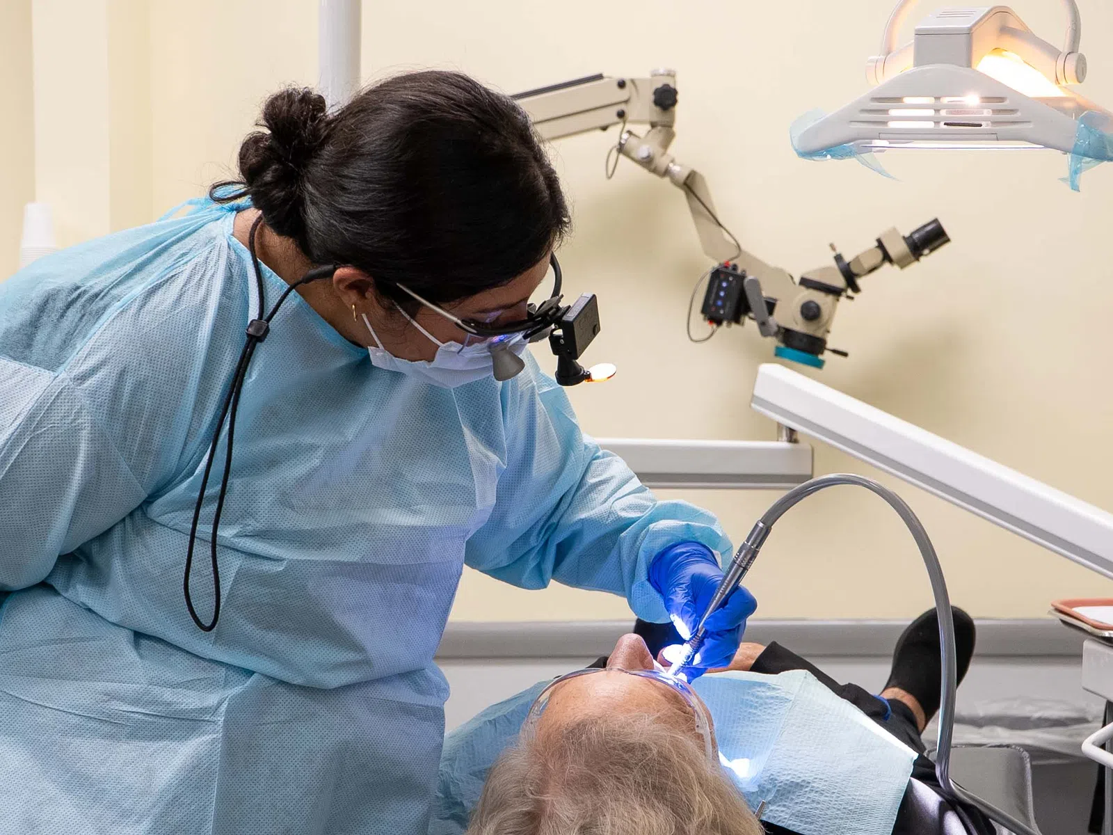Dentist in blue protective gear performing dental procedure on elderly patient's teeth with dental tools.