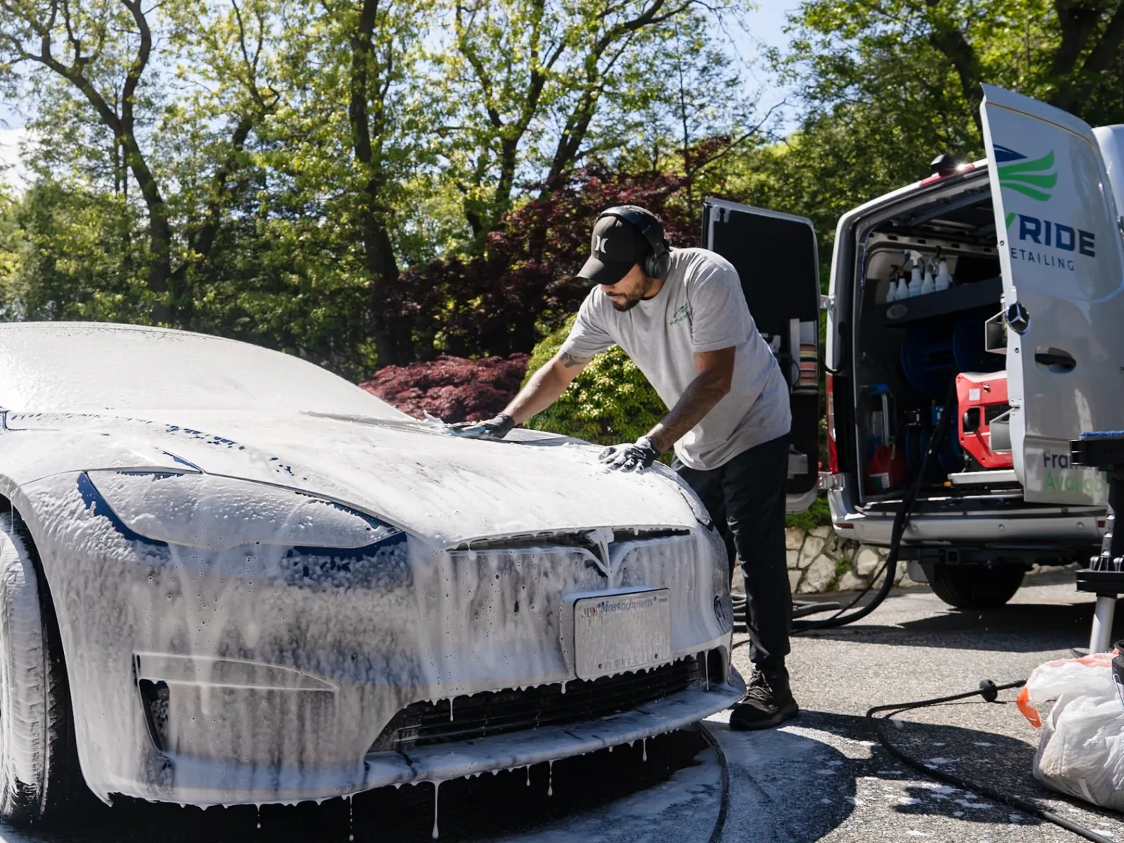 Man washing a white Tesla covered in foam with a cleaning van in a sunny outdoor setting.