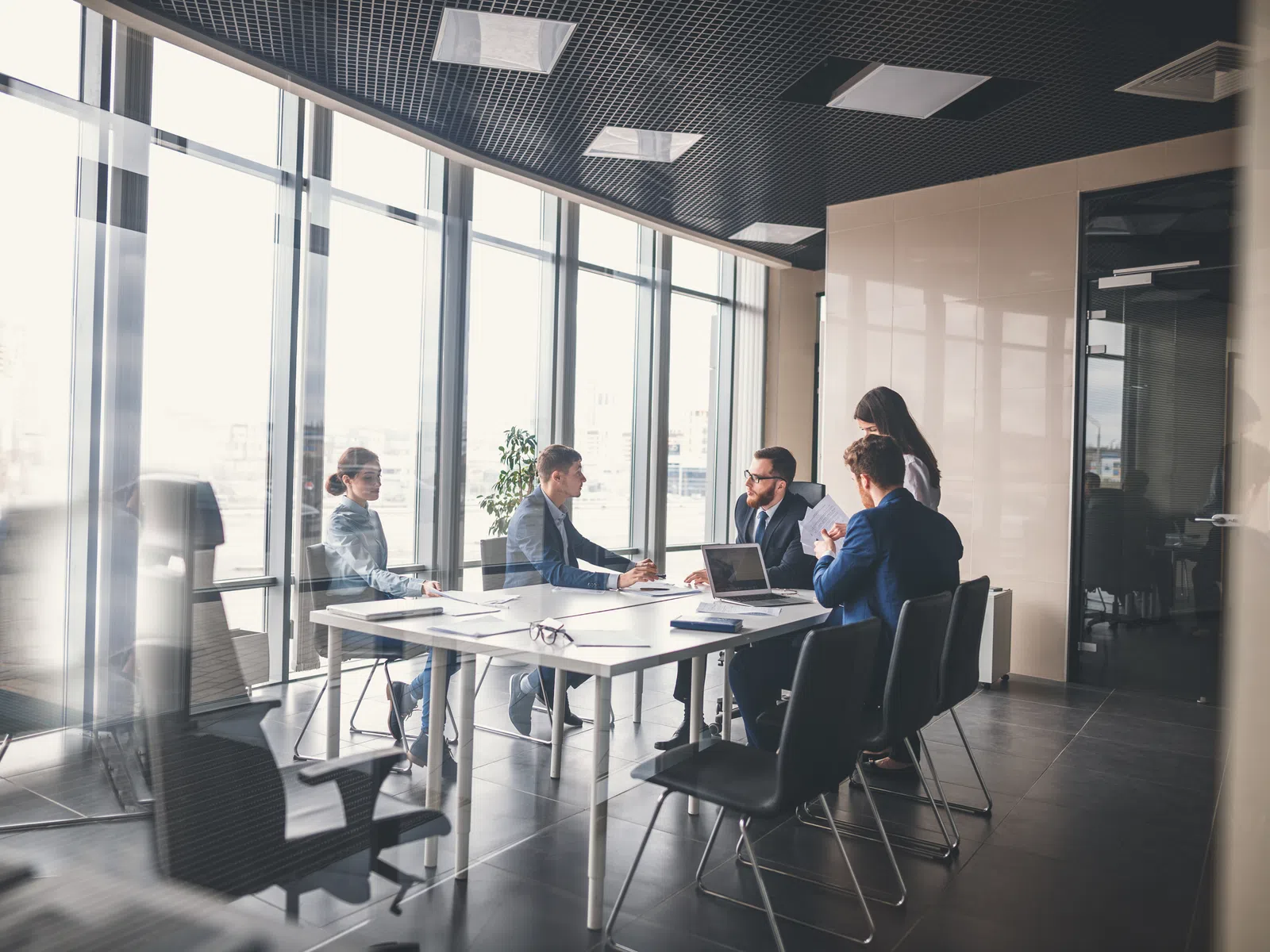 Business meeting with five professionals discussing ideas around a table in a modern office with large windows.